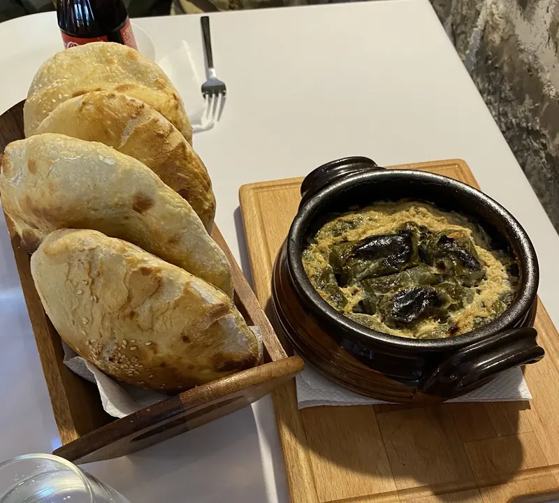 A baked dish in a clay pot next to warm bread pockets on a wooden board